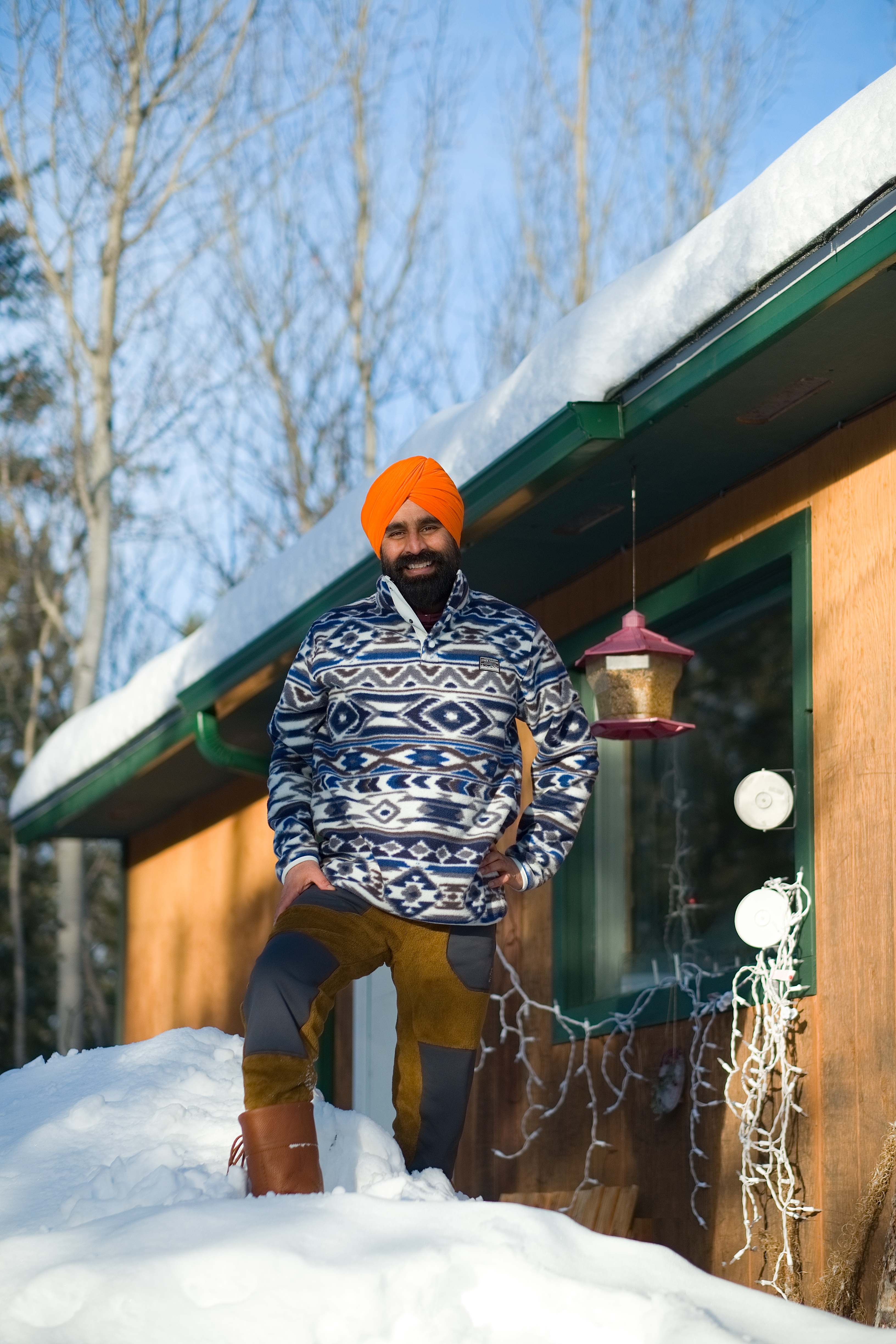 Photograph: Christian Kuntz Photography. Gurdeep stands outside a wooden walled cabin, that is covered in snow. He wears patterned knitwear and patchwork pants
